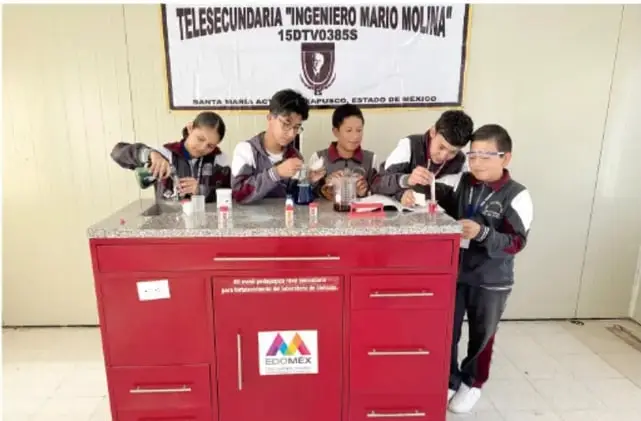 Fotografía de cinco estudiantes de la Telesecundaria "Ingeniero Mario Molina" (Santa Isabel, Estado de México) realizando prácticas de laboratorio en una mesa de trabajo roja con materiales de vidrio.