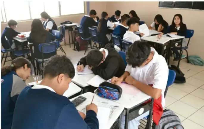 Fotografía de un grupo de estudiantes de Telesecundaria trabajando sentados en sus pupitres dentro del aula, algunos escribiendo en sus cuadernos.