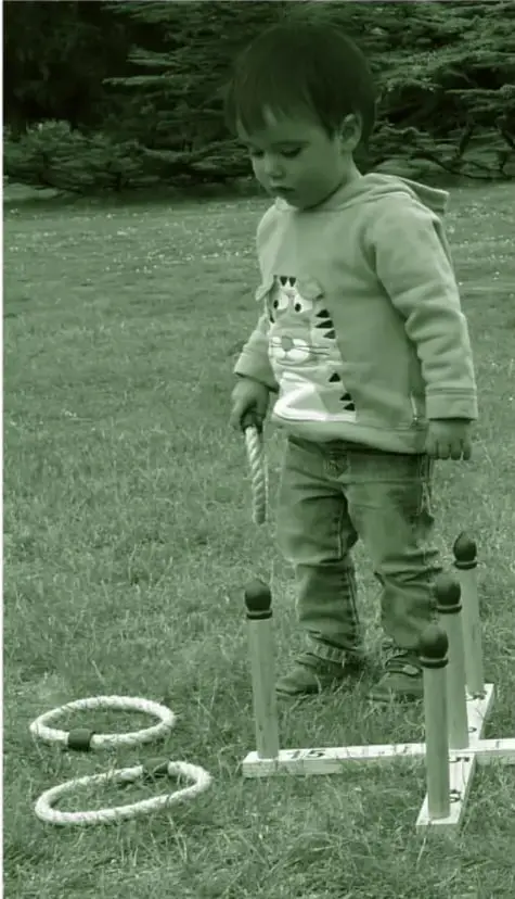 Fotografía en tonos verdes de un niño pequeño con sudadera y jeans, parado en el pasto, lanzando aros hacia un conjunto de estacas de madera; dos aros descansan en el suelo.