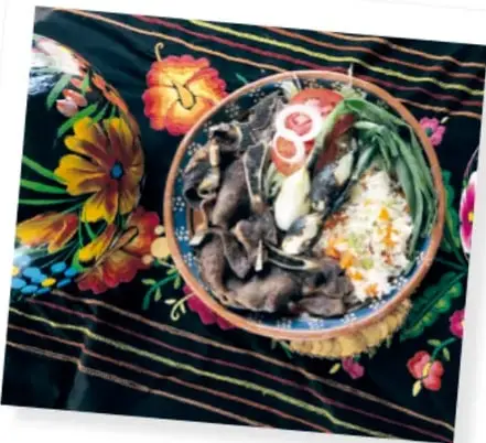 Fotografía de una ofrenda sobre un mantel negro bordado con flores de colores; se observa un plato de barro con comida tradicional y flores de cempasúchil.