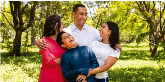 Fotografía de una familia (padre, madre e hijos) abrazándose y sonriendo en un parque, transmitiendo un ambiente de cuidado y protección familiar.