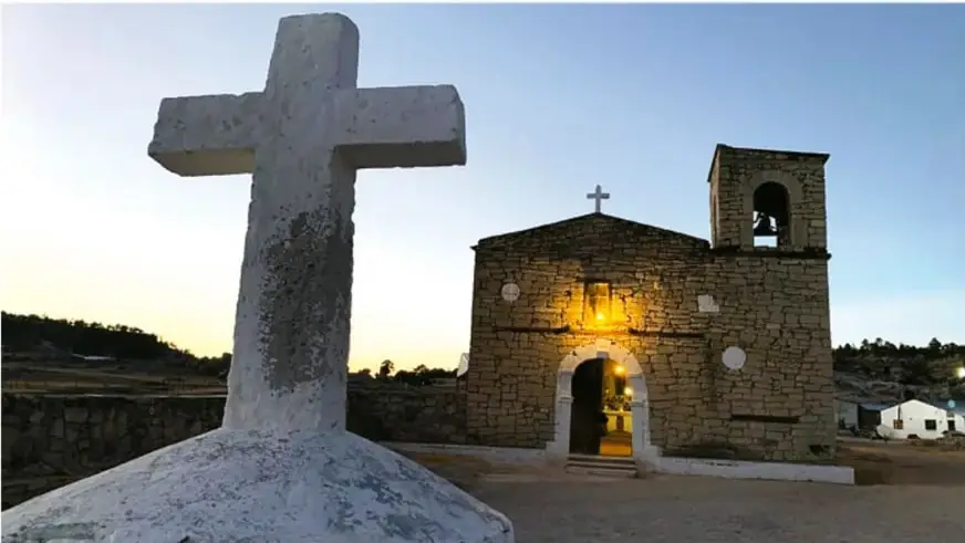 Fotografía de una misión religiosa de piedra al atardecer, con una gran cruz blanca en primer plano y una iglesia con campanario al fondo, iluminada por luz cálida.