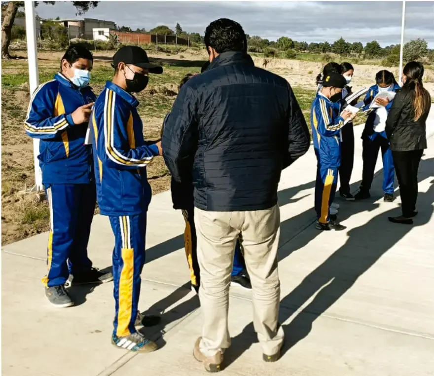 Fotografía de estudiantes de telesecundaria con uniforme azul y amarillo, junto con un adulto, en una actividad de campo en la que revisan documentos; crédito a Luis Eduardo García Medel, TOMATA.