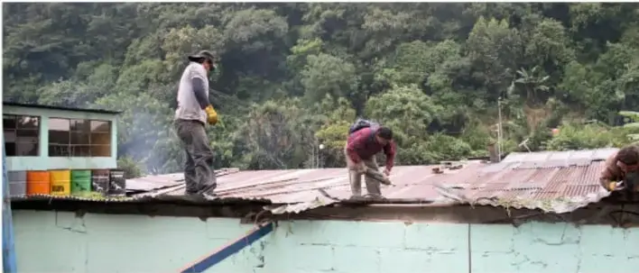 Fotografía de personas trabajando en el techo de una casa, asegurando láminas, en un entorno rural montañoso.