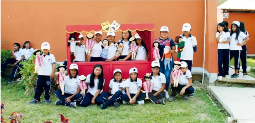Fotografía de un grupo de estudiantes de telesecundaria frente a un teatro de títeres rojo, con máscaras de comedia y tragedia colgando; algunos sostienen títeres.