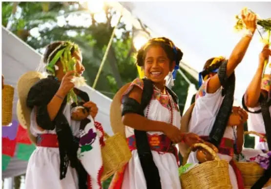 Fotografía de mujeres indígenas con vestimenta tradicional colorida, cargando canastas y ramas, en una celebración o actividad comunitaria al aire libre.