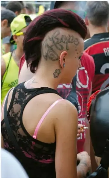 Fotografía de una persona joven de espaldas con el cabello rapado en los costados, tatuajes en la cabeza y vestido de encaje, en medio de una multitud, evocando estética contracultural.
