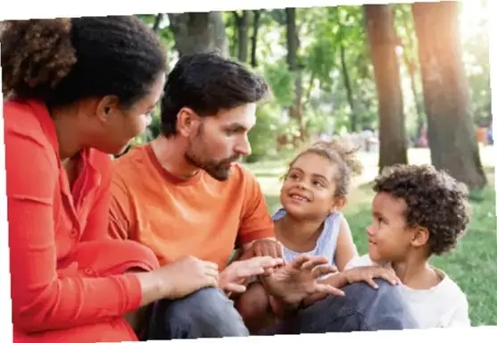 Fotografía de una familia (padre, madre y dos niños) sentada en el pasto conversando en un parque.