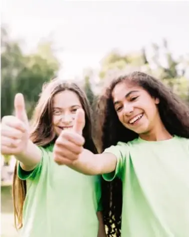 Fotografía de dos niñas sonriendo con gesto de pulgar arriba, vistiendo camisetas verdes, en un entorno al aire libre.