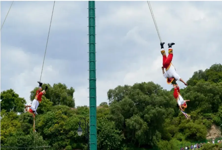 Fotografía de voladores: dos figuras humanas atadas por los pies a cuerdas que descienden girando desde lo alto de un mástil verde, con árboles y cielo nublado al fondo; representa una tradición o práctica cultural comunitaria.