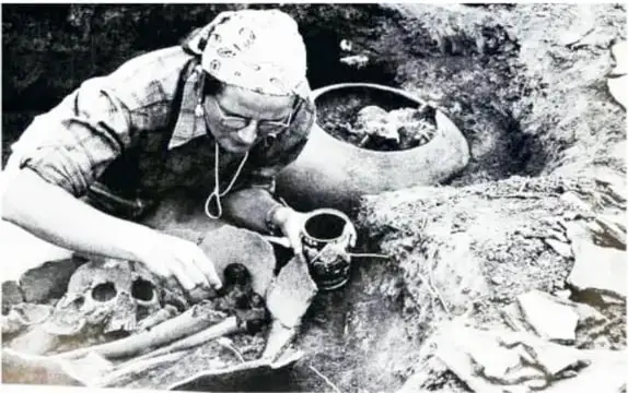 Fotografía en blanco y negro de la arqueóloga trabajando en la excavación, sosteniendo un cráneo humano junto a una vasija.