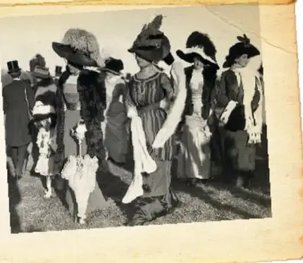 Fotografía antigua de mujeres adineradas caminando por la calle con vestidos largos, sombreros y sombrillas, acompañadas de niños.