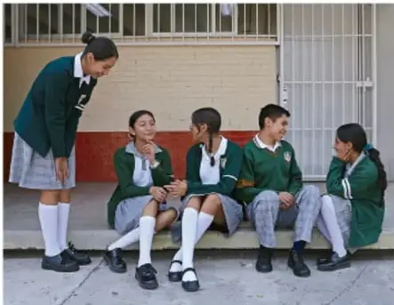 Grupo de estudiantes con uniforme escolar verde sentados en una banca al aire libre, conversando entre ellos