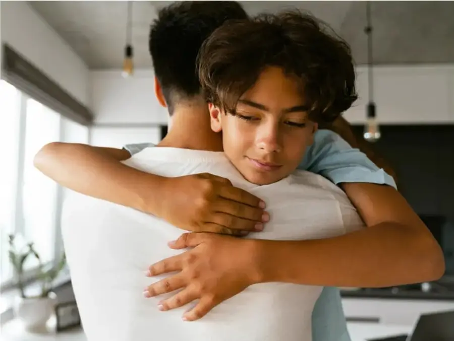 Fotografía de dos adolescentes abrazándose en un interior luminoso (cocina/sala), uno de espaldas con camiseta blanca y el otro de frente con camiseta azul, transmitiendo confianza y vínculo cercano.