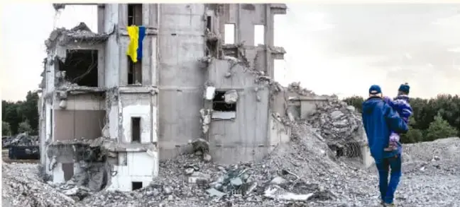 Fotografía de un edificio destruido por la guerra; en la fachada ondea una bandera de Ucrania (azul y amarillo) y dos personas con ropa de invierno caminan entre los escombros.