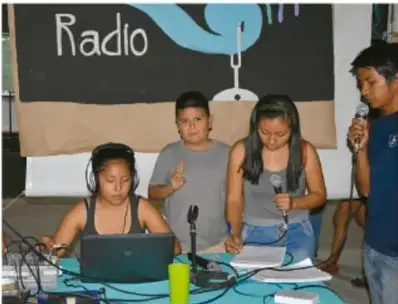 Fotografía de estudiantes de telesecundaria en un taller de radio escolar: cuatro alumnos con micrófonos y audífonos frente a una computadora, con un letrero que dice "Radio" en el pizarrón, ilustrando la expresión oral comunal.