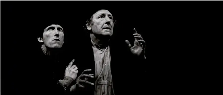 Fotografía en blanco y negro de una escena teatral con dos hombres (Vladimir y Estragón) en un escenario oscuro, uno de ellos mirando hacia arriba con expresión preocupada, ilustrando el diálogo de Esperando a Godot.
