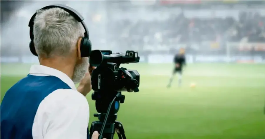 Fotografía de un camarógrafo con audífonos filmando un partido de fútbol desde una cámara profesional sobre trípode, en un estadio con aficionados de fondo.