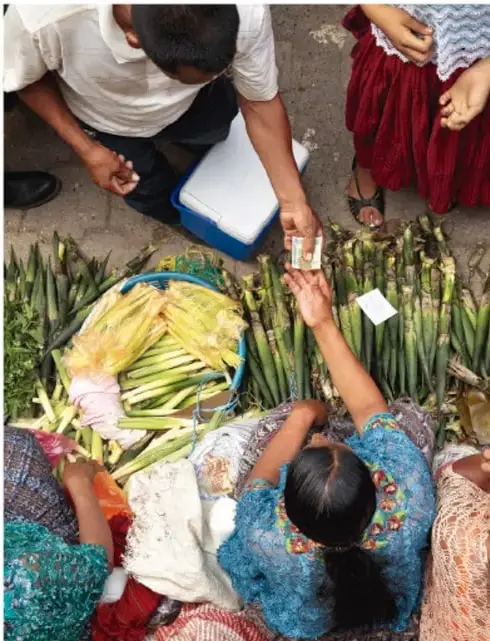 Personas sentadas en el suelo vendiendo o intercambiando alimentos como elotes y verduras; una persona recibe dinero.