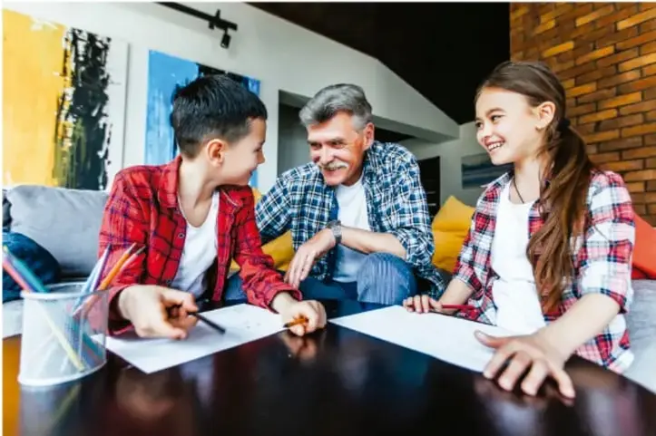 Fotografía de dos niños y un adulto mayor sentados a una mesa; los niños toman notas en hojas blancas mientras conversan con el abuelo, que sonríe. Representa la entrevista a una persona mayor.