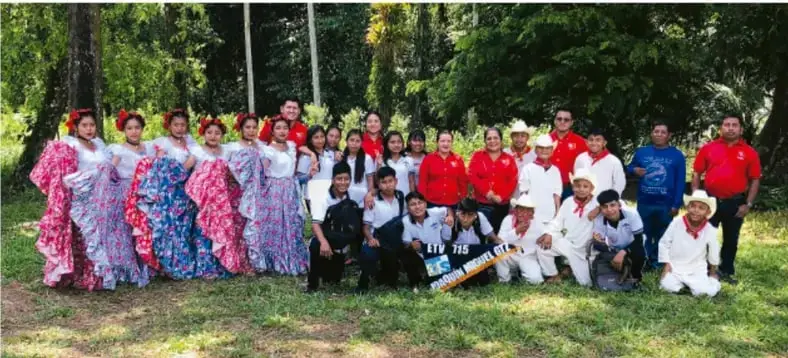 Fotografía de un grupo de personas jóvenes y adultas al aire libre, entre árboles; varias mujeres con vestidos de folclor de colores, hombres con camisas y sombreros, una persona con instrumento musical de viento, posando para la foto.