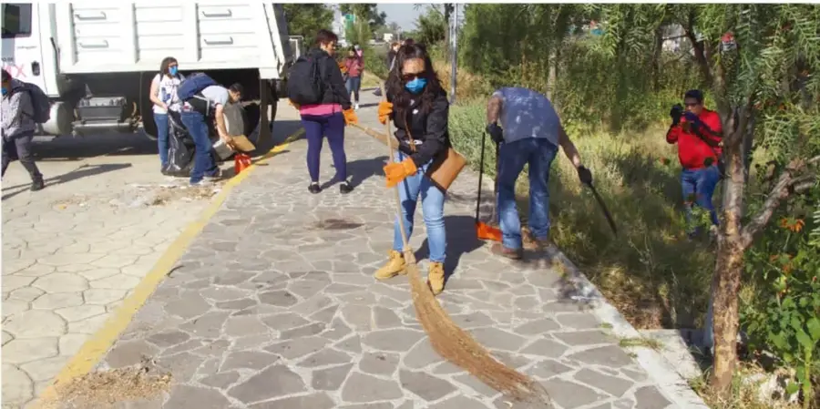 Fotografía de estudiantes y adultos con cubrebocas barriendo y recogiendo basura en una calle pavimentada, junto a un camión de carga; se observa trabajo comunitario de limpieza.