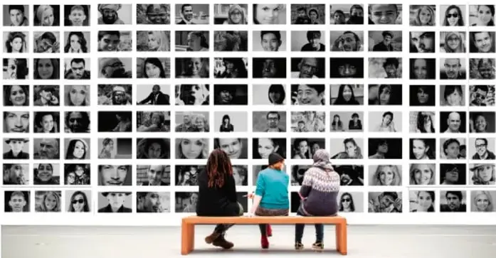Mosaico de muchos retratos fotográficos en blanco y negro de personas de distintas edades y contextos; al frente, tres niñas sentadas en una banca observan el mosaico, símbolo de la diversidad.