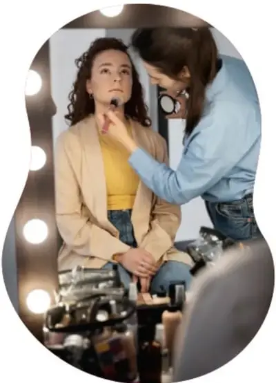 Fotografía circular que muestra a una persona siendo maquillada frente a un espejo con luces de camerino, ilustrando la preparación para una puesta en escena teatral.