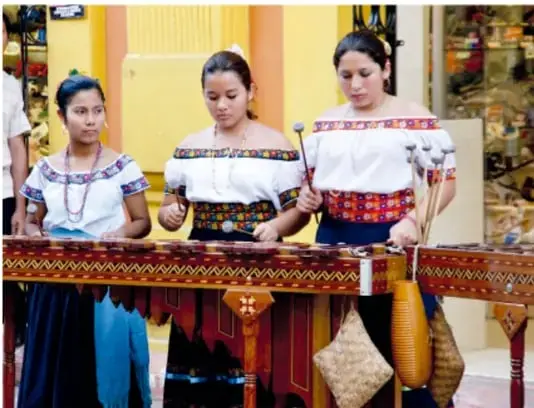 Tres niñas con blusas bordadas tocando una marimba de madera con mazos.