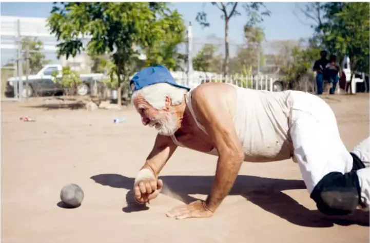 Hombre mayor con gorra azul y camiseta blanca lanzándose en una cancha de tierra para golpear una pelota de hule; al fondo, árboles y vehículos.