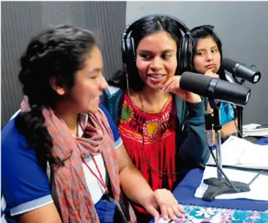 Fotografía de tres mujeres jóvenes en un estudio de radio, con audífonos y micrófonos, participando en la grabación de un programa.