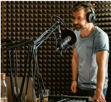 Fotografía de un hombre con audífonos frente a un micrófono con cortavientos en un estudio con paneles acústicos, ilustrando el trabajo de sonido y doblaje.