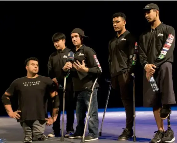 Fotografía de un grupo de cinco jóvenes en un escenario oscuro; uno habla con micrófono, otros usan muletas o prótesis, vestidos con ropa negra deportiva; aparenta ser una presentación o actividad inclusiva.