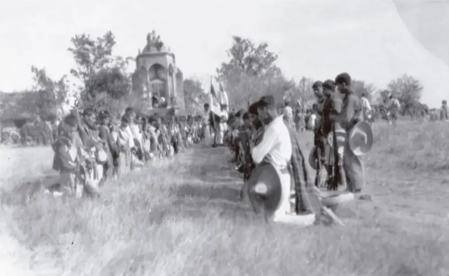 Fotografía histórica en blanco y negro que muestra una asamblea o ceremonia al aire libre: grupos de personas alineadas, algunas con sombreros y portando objetos, frente a un monumento o altar con figuras, en un entorno campestre. Ilustra prácticas y vida social de la posrevolución mexicana.