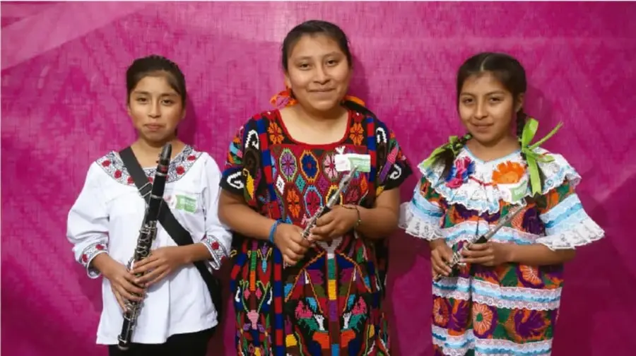 Fotografía de tres niñas y una mujer de pie frente a un fondo rosa, con trajes tradicionales indígenas bordados y portando instrumentos musicales (clarinete y flauta); se les identifica como participantes de una actividad cultural.