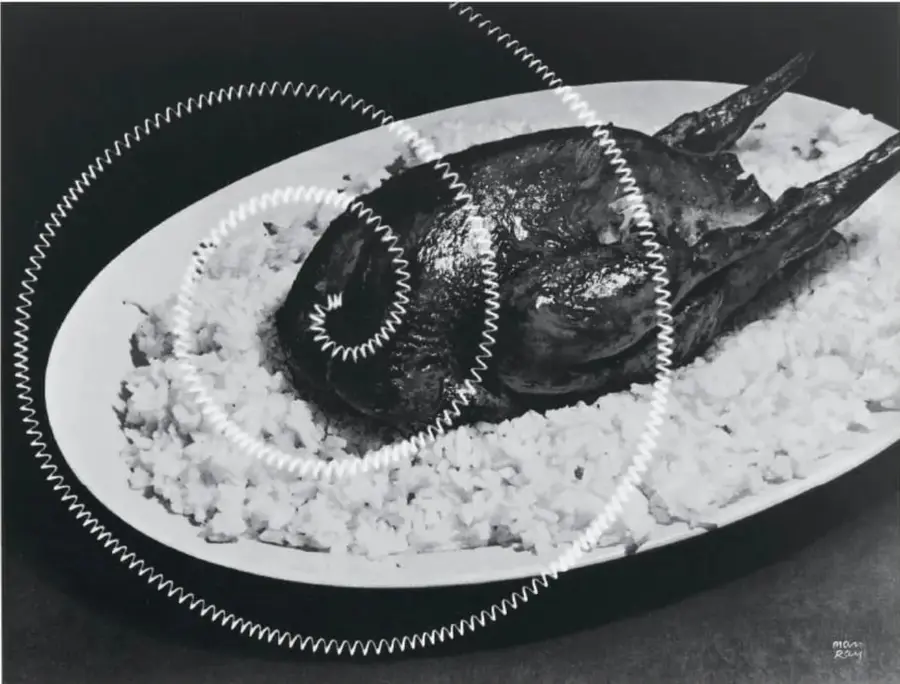 Fotografía en blanco y negro de un plato ovalado con arroz y un ave asada, rodeada por una espiral de alambre blanco. Obra de Man Ray.