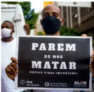 Fotografía de una persona con cubrebocas negro sosteniendo un cartel que dice "PAREM DE NOS MATAR - NOSSAS VIDAS IMPORTAM!" (¡Paren de matarnos, nuestras vidas importan!), protesta en portugués contra el racismo.