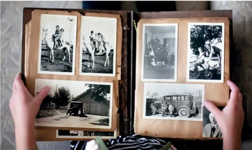 Manos sosteniendo un álbum antiguo abierto con fotografías en blanco y negro de personas, caballos, un automóvil y una casa, como ejemplo de álbum comunitario.