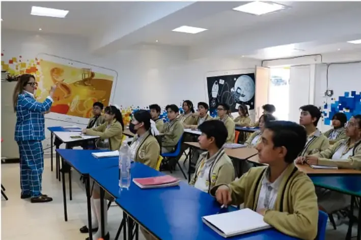 Fotografía de un aula de telesecundaria donde una docente frente a una pantalla digital explica a los estudiantes, quienes están sentados en mesas azules tomando notas; en la pared hay murales y posters educativos.