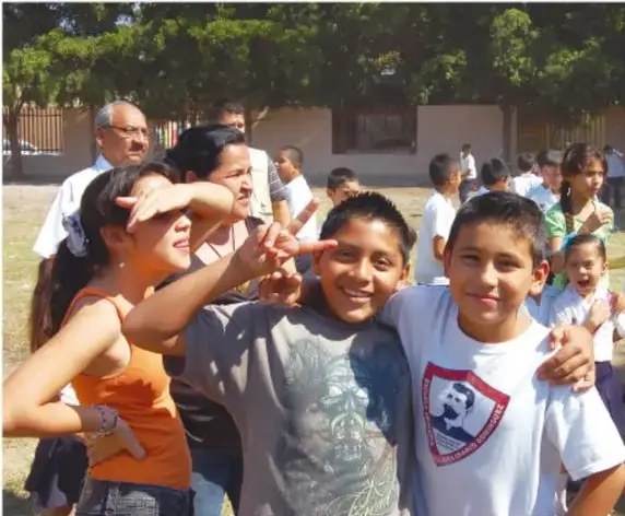Fotografía de estudiantes de primaria al aire libre, sonriendo y haciendo señas con las manos, acompañados de algunos adultos al fondo.