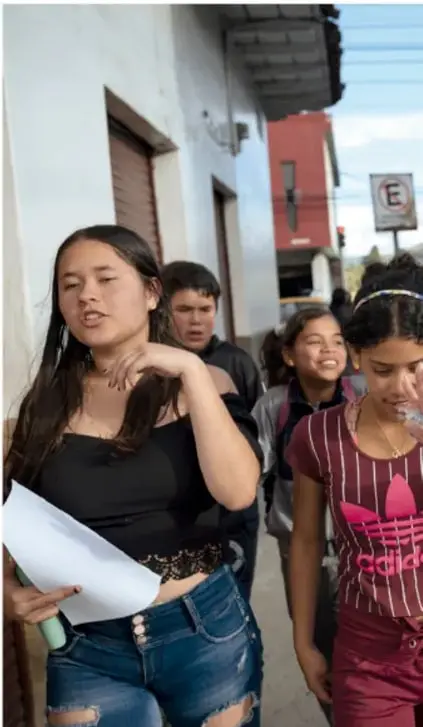 Fotografía de un grupo de adolescentes caminando por una calle; una joven al frente habla o gesticula mientras sostiene un papel, acompañada de otros estudiantes, ilustrando la interacción social cotidiana.