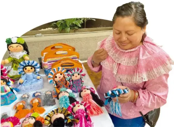 Mujer indígena con blusa rosa bordada sostiene una muñeca Lele mientras exhibe varias muñecas sobre una mesa.