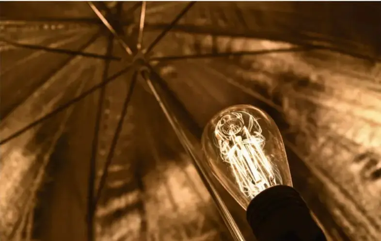 Fotografía en tonos sepia de una bombilla eléctrica encendida con filamento visible, iluminando la estructura interna de una cúpula o sombrilla, evocando la innovación de Thomas Alva Edison.