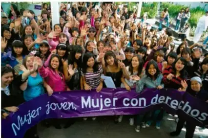 Asamblea de personas sentadas en mesas redondas, una mujer de pie habla con un micrófono.