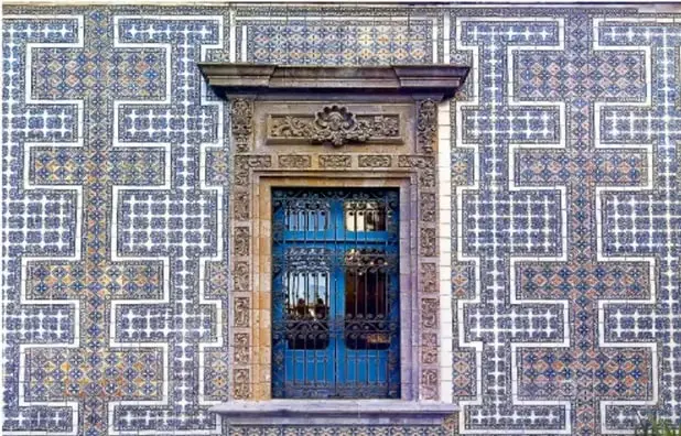 Fachada de un edificio con azulejos azules y blancos formando un patrón geométrico teselado, con una puerta de hierro forjado en el centro.