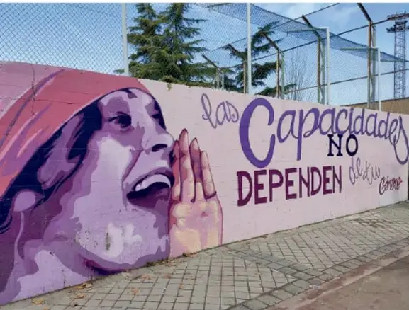 Mural urbano en una pared con el rostro de una persona con hidrocefalia (mujer con gorro rosa) y la frase 'Las capacidades NO dependen de nadie', junto a una cancha deportiva con malla.
