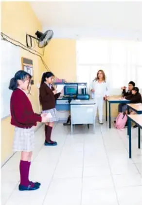 Fotografía de estudiantes en un salón de clase con equipo de cómputo, uno de ellos de pie frente a sus compañeros.