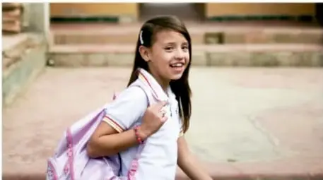 Fotografía de una niña con mochila rosa y uniforme escolar caminando y sonriendo en un patio escolar.