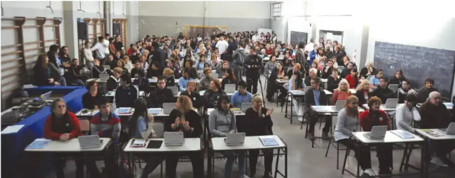 Fotografía de un aula con muchos estudiantes sentados en mesas, algunos con computadoras portátiles, y un pizarrón al fondo.