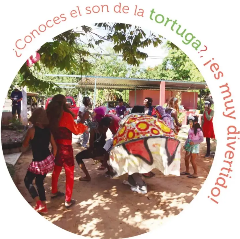 Personas bailando y mirando alrededor de una figura vestida de tortuga con un caparazón grande y decorado con círculos de colores, en un patio de tierra con árboles y casas.